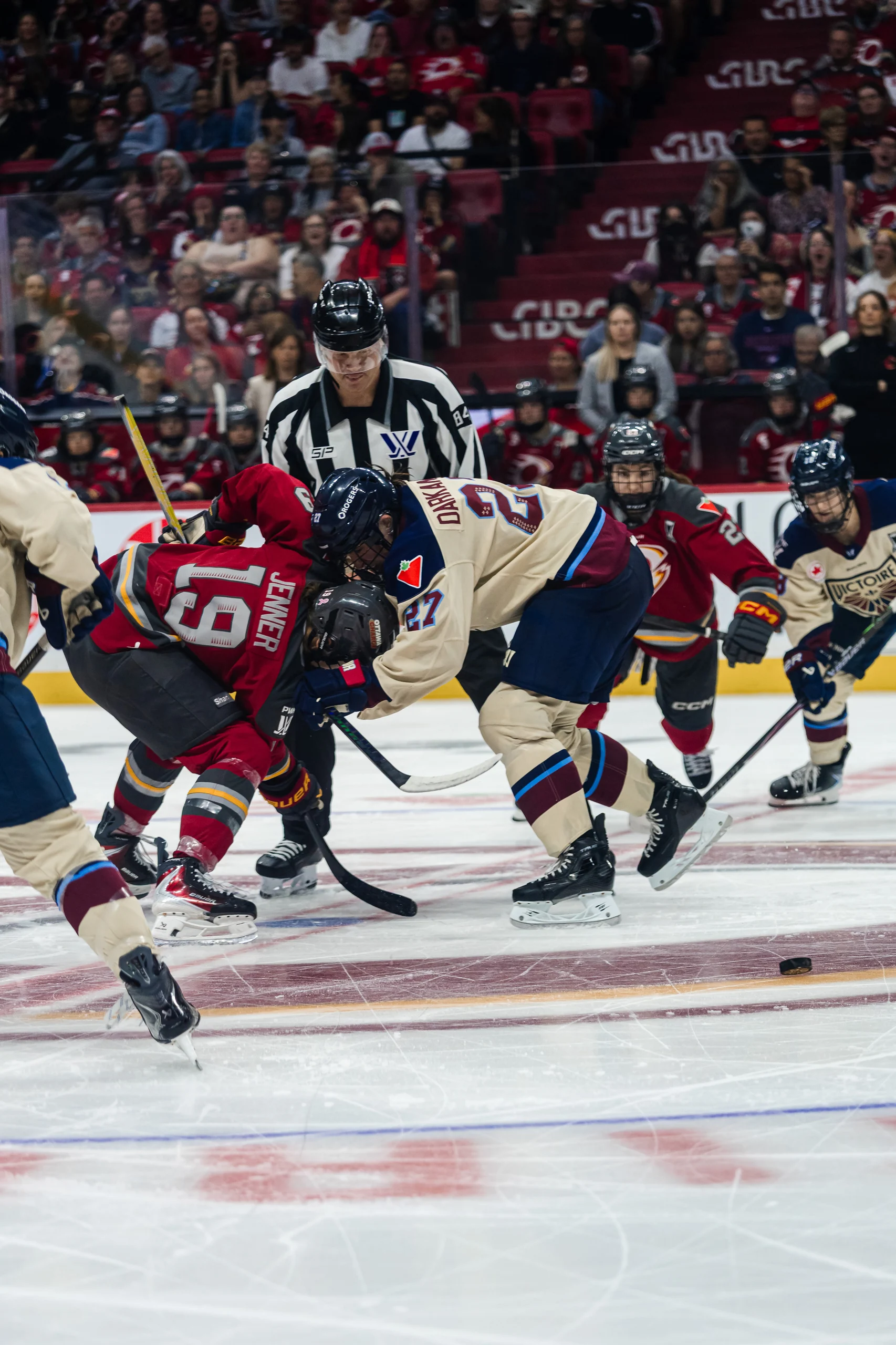 A faceoff between an Ottawa Charge forward in red and a Montréal Victoire forward in cream, the official watching over them.