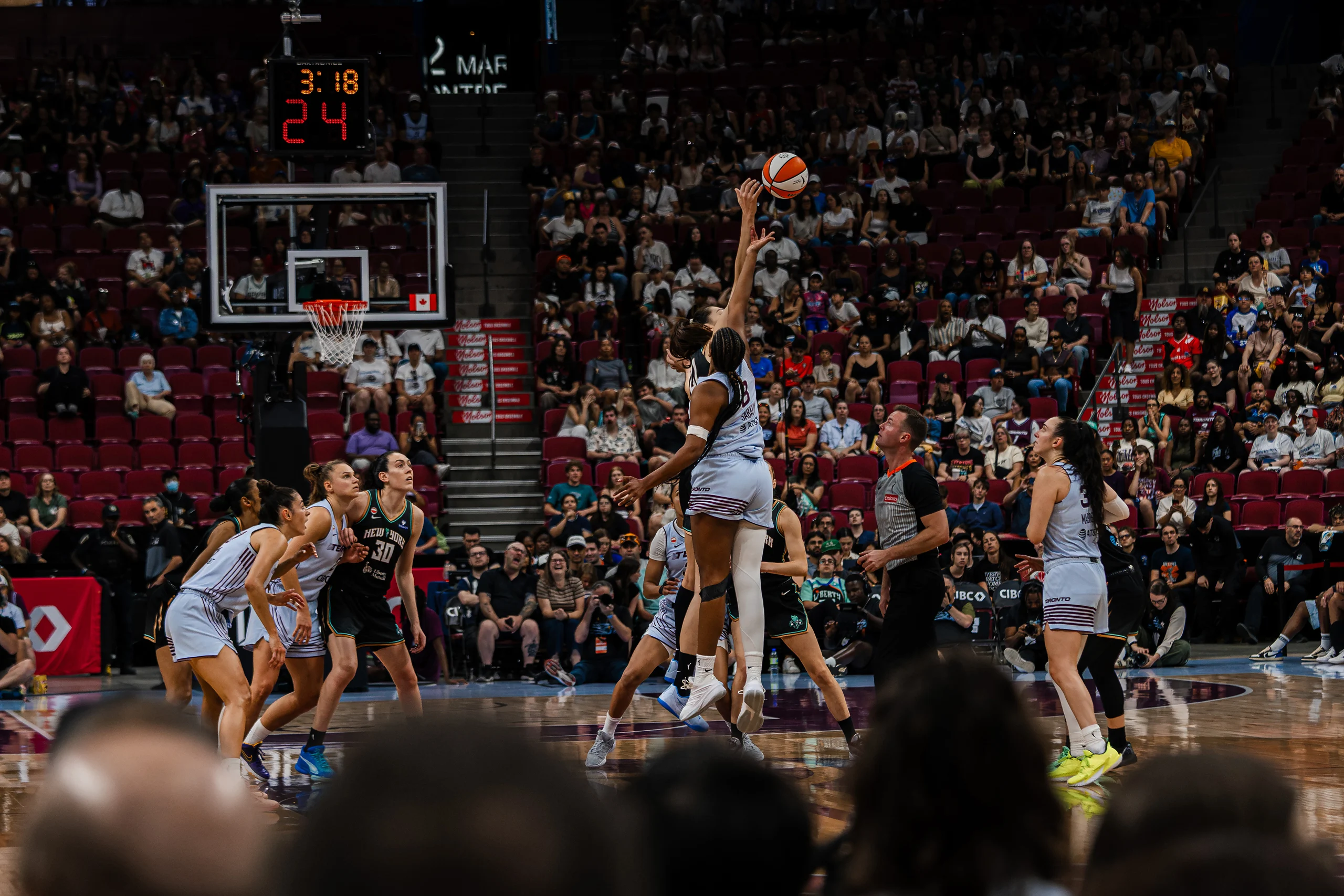 Two players stretch for the opening tip-off, the ball just above their fingertips, in front of a full arena.
